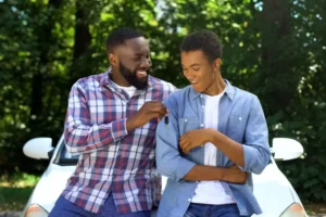 father and son sitting on the hood of a car. The dad is giving his son the keys to the car