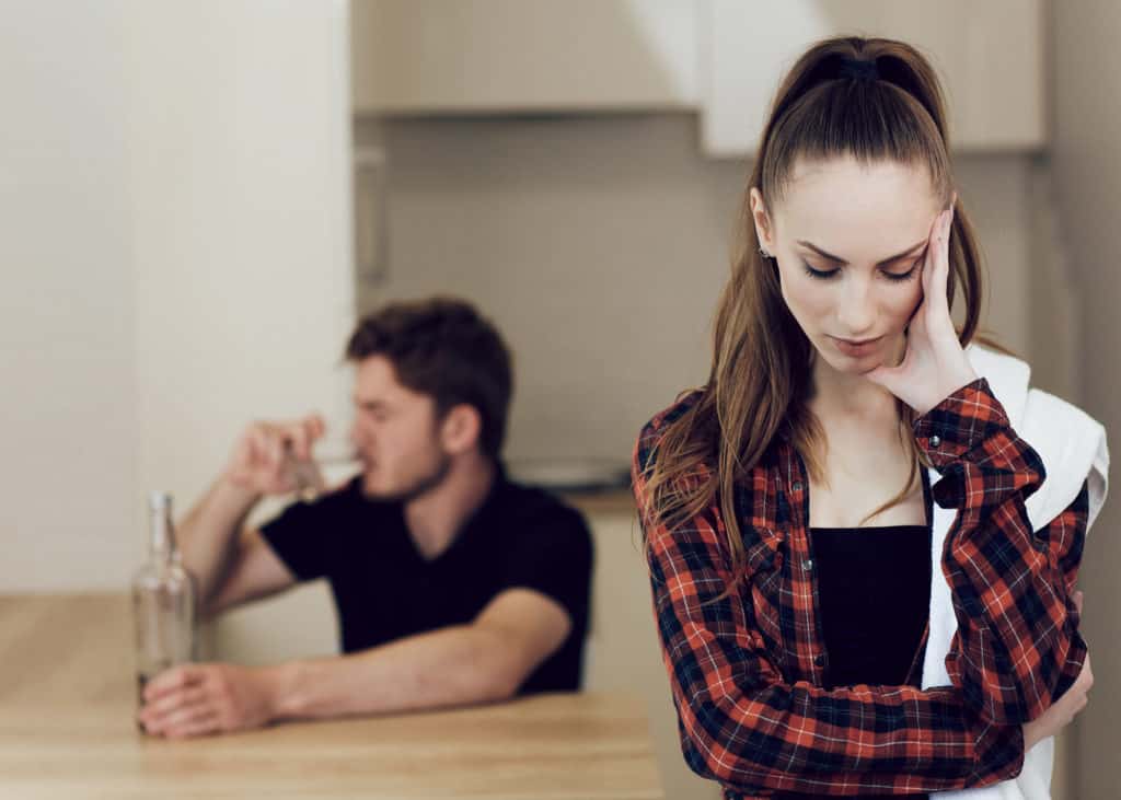 Upset-woman-who-is-spouse-of-an-addict-with-drunk-husband-in-background