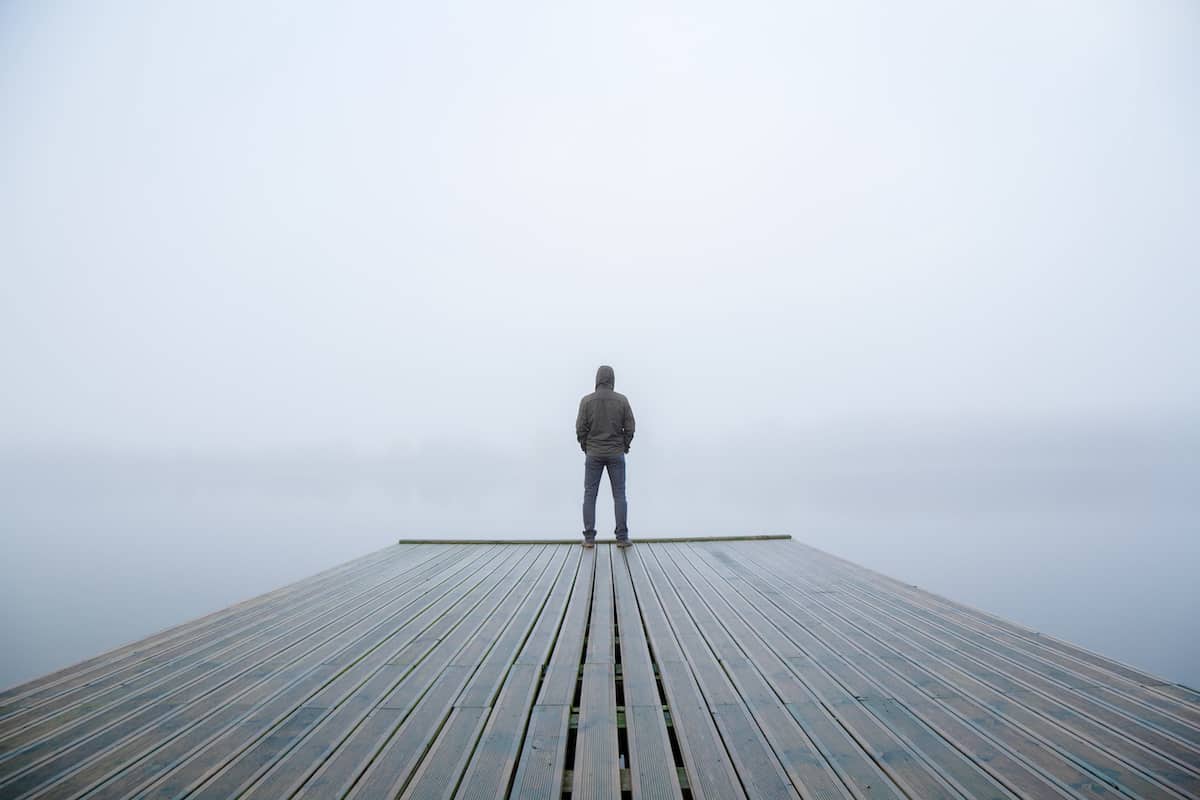 Young man standing alone on edge of footbridge and staring at lake. Mist over water. Foggy air. Early chilly morning in autumn. Beautiful freedom moment and peaceful atmosphere in nature. Back view.