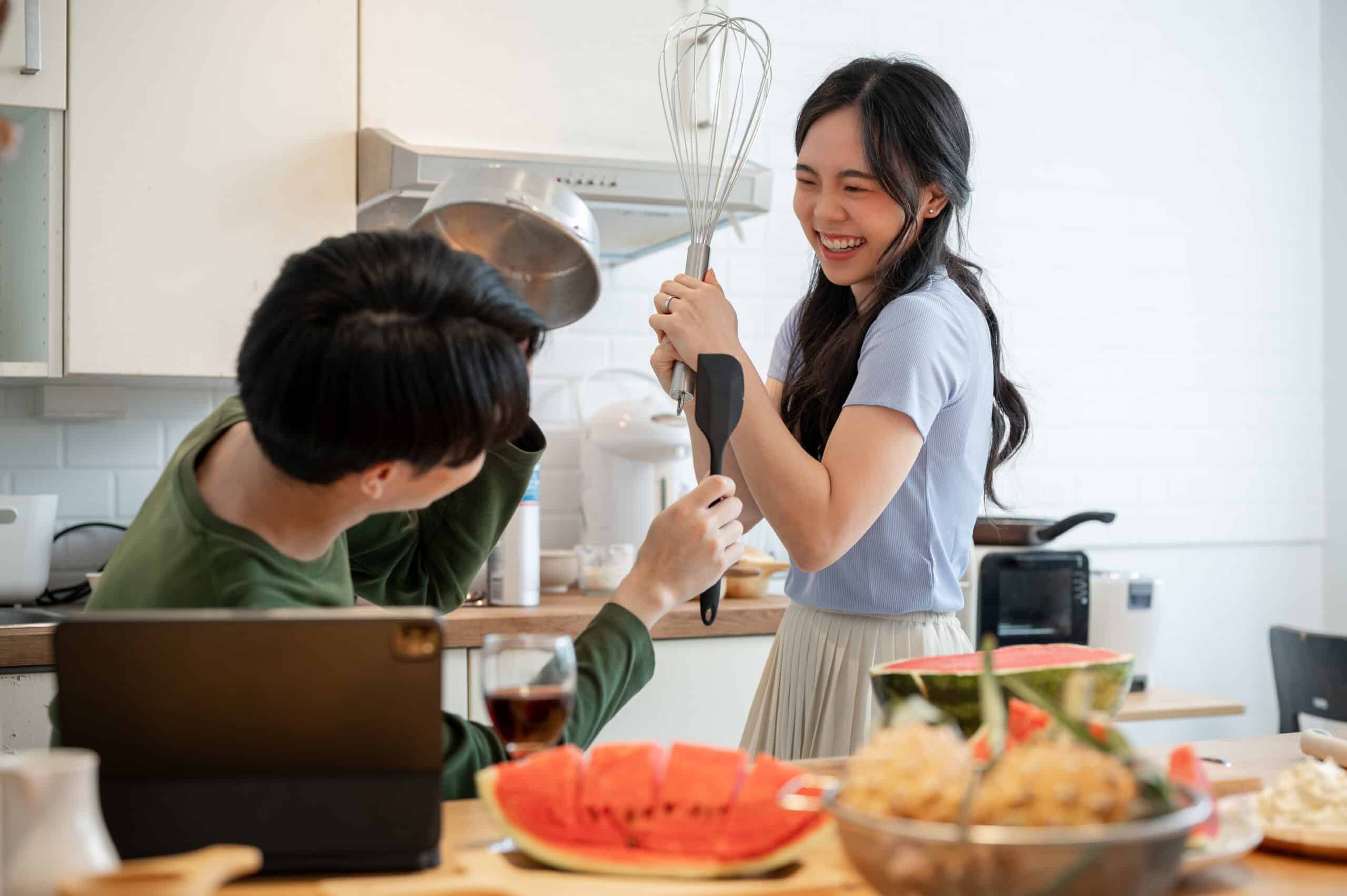A lovely and playful young Asian couple is pretending fight with utensils tools and having fun while preparing food in the kitchen together. Fun domestic life, cooking date, married couple at home